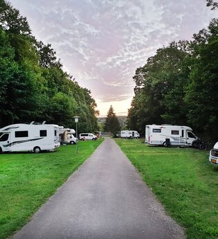 Blick auf den Wohnmobilstellplatz beim Naturcamping in Bad Mergentheim. Die Wohnmobile stehen auf Grünflächen. Die Bäume rechts und links sind groß und intensiv grün. Die Wolken am Himmel sind rosafarben. Im Hintergrund geht gleich die Sonne auf/unter. | © Unbekannt