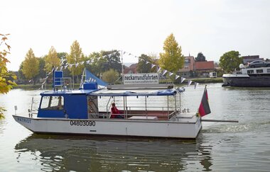 Ein Flug mit einem kleineren weiß-blauen Motorboot mit einer Wimpelkette. Im Hintergrund ein Dorf mit Bäumen. Haßmersheim im Odenwald. | © Touristikgemeinschaft Odenwald e.V.