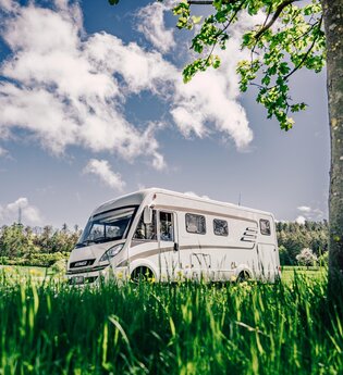 Weißes Wohnmobil fährt auf einer Straße. Im Vordergrund ist eine Blumenwiese im Hintergrund eine Streuobstwiese mit angrenzendem Wald. | Odenwald | © Touristikgemeinschaft Odenwald e.V.