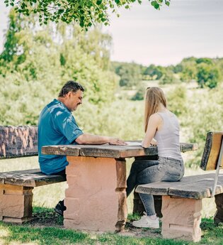 Ein mann und eine Frau sitzen an einem Sitzgelegenheit aus Stein. Im Hintergrund sind Wiesen und Felder. Mudau im Odenwald. | © Touristikgemeinschaft Odenwald e.V.