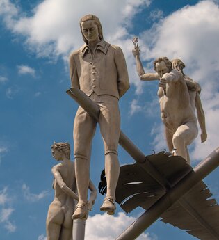 Am Ortseingang von Lauffen am Neckar erhebt sich das eindrucksvolle Kunstwerk „Hölderlin im Kreisverkehr“ des Bildhauers Peter Lenk. Auf einem geschwungenen „H“ balanciert eine überdimensionale Schreibfeder, auf der Friedrich Hölderlin als Kind und als ju | © Stadt Lauffen am Neckar