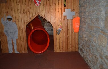 Blick auf einen Rutschenausgang in der Spielscheune in Röttingen. Der Ausgang ist mit einer Holzwand verkleidet. Ein gemalter Ritter hält daneben Wache. | © Tourismusverband "Liebliches Taubertal" e.V.