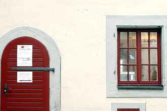 Eingangstüre zur Touristinformation aus dunkelrot gestrichenem Holz, rechts daneben zwei, dunkelrot gestrichene Sprossenfenstertüre, | © Petra Natzkowski